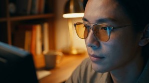 Portrait rapproché d'une personne portant des lunettes à verres teintés devant un écran d'ordinateur le soir.